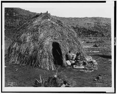 A dome-shaped hut made of brush and grass stands in a barren landscape, with several baskets and pottery placed near its entrance.