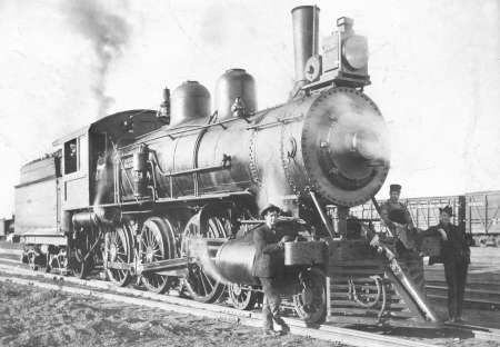 Black and white photo of a steam locomotive on tracks, with two men standing beside it; one is leaning against the front wheel.