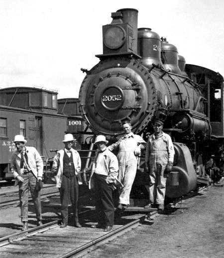 Five men stand in front of a steam locomotive numbered 2052, with train cars visible in the background on a rail yard.
