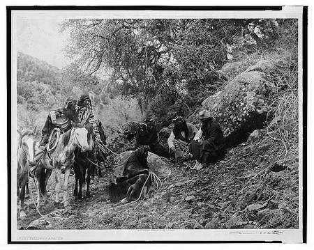 A group of Apache people sit and stand near horses on a rocky, wooded hillside, with trees and boulders surrounding them.