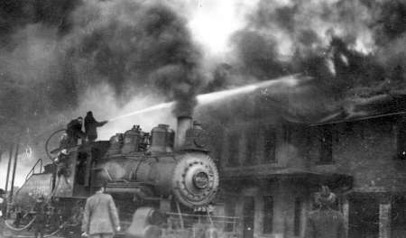 A steam locomotive sprays water onto a burning building as thick smoke rises; several people are present near the scene.