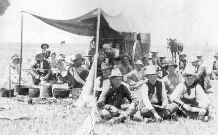 A group of cowboys sit and eat around a chuckwagon under a canopy on an open prairie, with cooking pots and horses visible in the background.