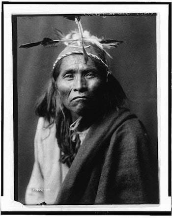Black-and-white portrait of an Indigenous man wearing a feathered headdress and a blanket, looking directly at the camera.