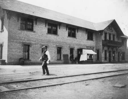 Black-and-white photo of a man walking across railroad tracks in front of a two-story brick train station building with several people near the entrance.