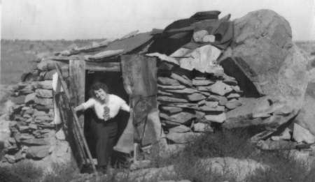 A woman stands in the doorway of a small, makeshift stone and wood shelter in a rural landscape.