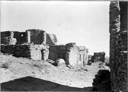 Black and white photo of old stone ruins on a dusty, uneven ground under a clear sky; partial shadow in the foreground, with crumbling walls and doorways visible.