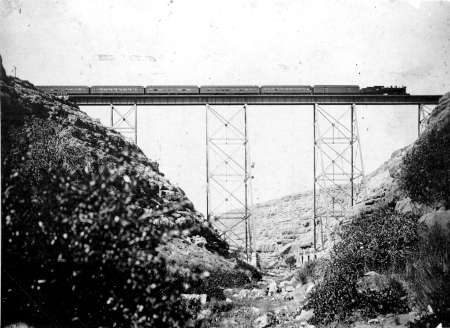A train crosses a tall, narrow steel bridge spanning a rocky valley with sparse vegetation.