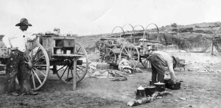 Two people prepare food at a chuckwagon camp, with cooking pots over a fire and covered wagons in the background on open terrain.