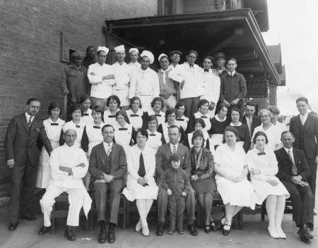 A large group of men and women, some in uniforms and some in formal attire, pose together in front of a brick building for a formal group photo.