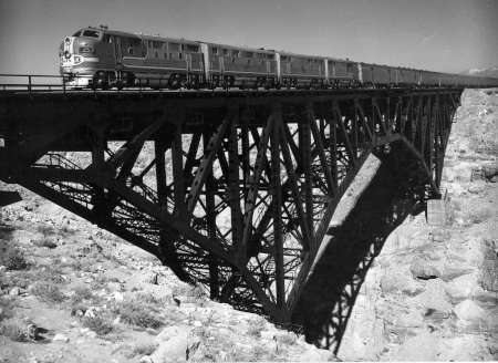 A long train with multiple cars crosses a tall steel bridge over a rocky canyon, with shadows cast by the bridge’s structure.