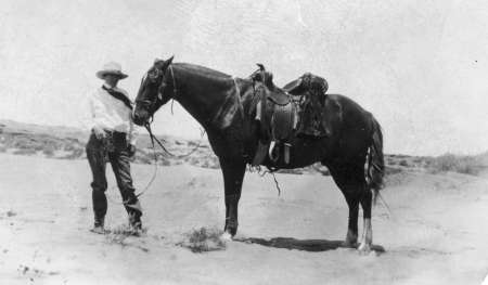 A man in a hat stands in the desert holding the reins of a saddled horse.