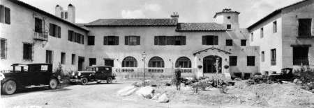 Black-and-white photo of a large two-story Spanish-style building with arched windows, vintage cars parked in front, and a landscaped entrance.