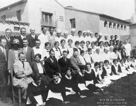 A large group of men and women, some in uniforms and aprons, pose for a formal group photo outside a stucco building.