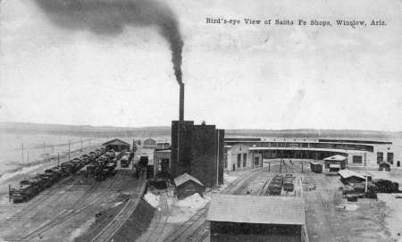 Black-and-white photo of Santa Fe Shops in Winslow, Arizona, showing trains, tracks, and a large building with a tall smokestack emitting smoke.
