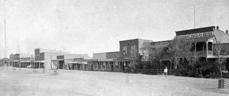 Black and white photo of a wide dirt street lined with old western-style buildings, including a hotel; a few people and trees are visible.