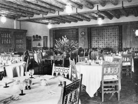 A historic dining room with wooden beam ceiling, decorated chairs, set tables with linens and glassware, and a large floral arrangement at the center.