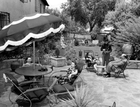 Several people relax on lounge chairs and around a table under a large umbrella in a landscaped backyard on a sunny day.