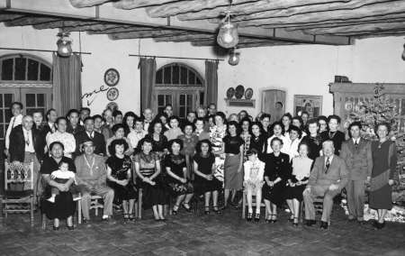 Large group of men and women pose indoors for a formal photo, some seated and others standing, in a room with wooden ceiling beams and decorative plates on the walls.