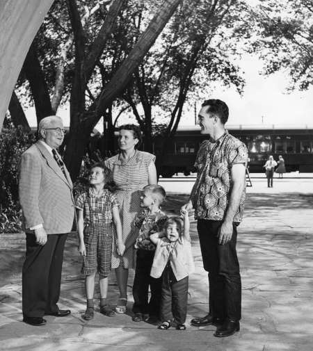 A group of five people, including three children and two adults, stand outdoors talking to an older man in a suit. A train and trees are visible in the background.