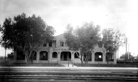 Black-and-white photo of a two-story Santa Fe railroad depot building, partially obscured by trees, with train tracks in the foreground.