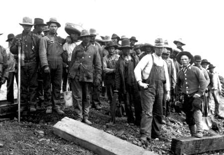 A group of men wearing work clothes and hats stand together on rocky ground, some holding tools, in a black and white historical photograph.