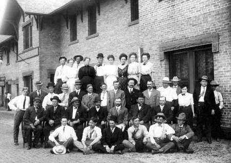 A black and white photo of a large group of men and women posing in rows outside a brick building. Most are wearing formal attire typical of the early 20th century.