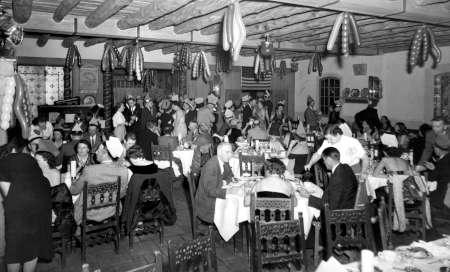 Black and white photo of a crowded restaurant or banquet hall with people dining at tables and decorations hanging from the ceiling.