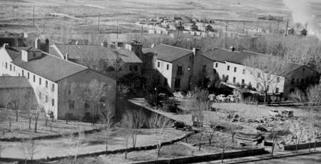 Black-and-white aerial photo of several large buildings surrounded by bare trees and open land, with smoke rising from a distant area in the background.