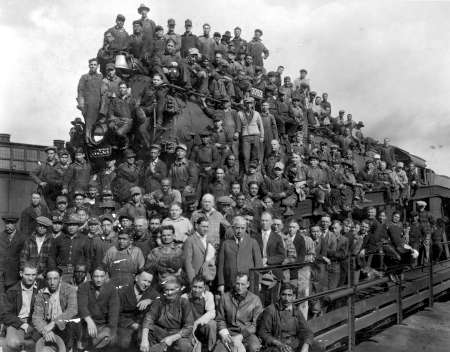 A large group of men pose together on and in front of a train engine at an industrial site, many wearing work uniforms and hats, with a few in suits.