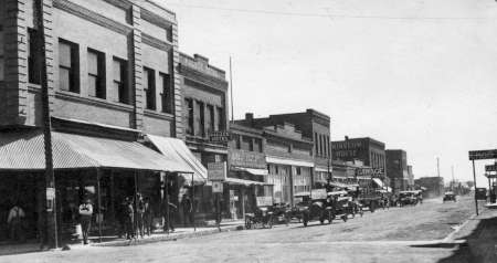 Black and white photo of a small town main street lined with early 20th-century buildings, shops, and parked vintage automobiles. Pedestrians stand on the sidewalk.