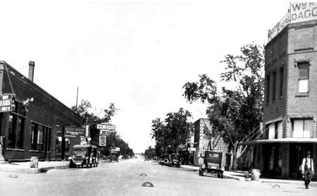 Black and white photo of a small town street with early automobiles parked along the sides and various business signs visible, including "Curios" and "Barber Shop.