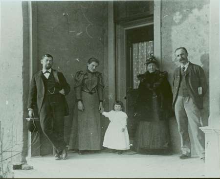 Five people, including a child in a white dress, pose on a porch in front of a doorway; the adults are wearing late 19th-century clothing.