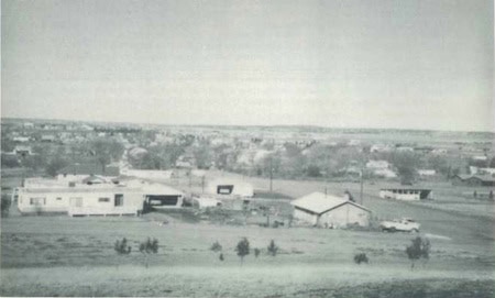 Black and white photo of a rural area with several buildings, houses, and open fields, with a town visible in the background.