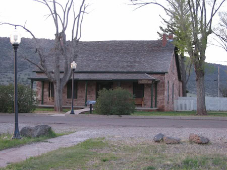 A single-story brick house with a covered porch sits behind a street, surrounded by a few trees and a lamppost, with hills in the background.