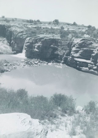 A small waterfall flows over rocky cliffs into a pool below, surrounded by vegetation and shrub-covered terrain under a clear sky.