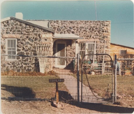 A small stone house with a fenced front yard, a narrow sidewalk leading to the door, and a sign reading "Coplan" near the gate.