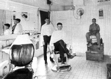 Black and white photo of a barber standing next to a man in a barber chair, with another man sitting on a shoeshine stand in a vintage barbershop.