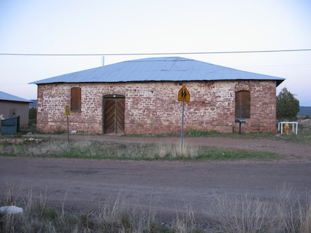 A weathered brick building with a metal roof sits beside a rural road, flanked by two pedestrian crossing signs and sparse vegetation.