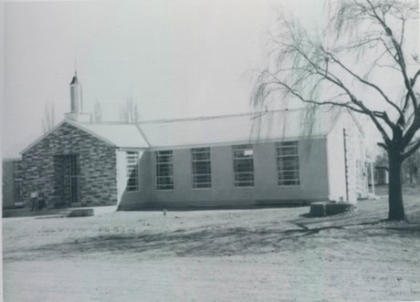 Black and white photo of a one-story building with a small steeple, brick facade, several windows, and a leafless tree in the foreground.