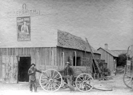 Two men stand beside a large wooden wagon wheel in front of a blacksmith shop made of wood with a shingled roof; other wagons and buildings are visible nearby.