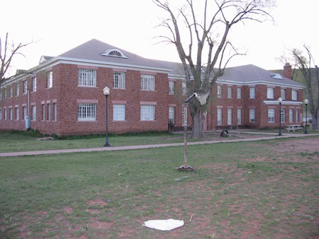 A two-story red brick building with white-trimmed windows, barred on the lower level, stands behind a patchy lawn and path with a few leafless trees.
