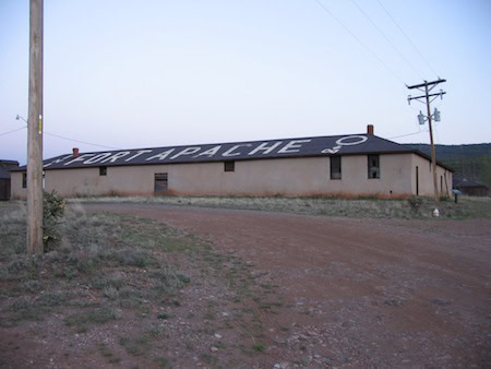 A long, single-story building with "FORT APACHE" painted on the roof, situated on a dirt road with utility poles and sparse grass in the foreground.