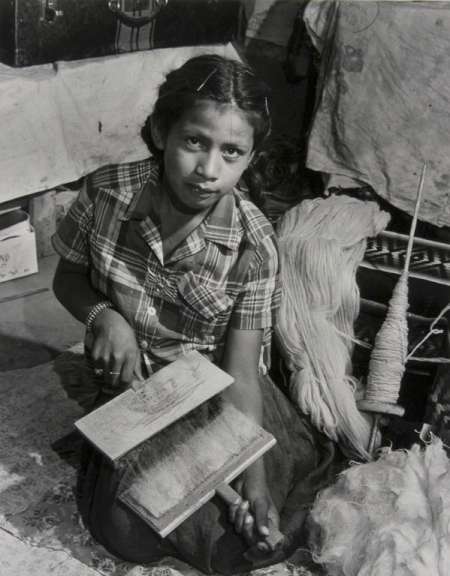 A woman sits on the floor, carding fibers with hand carders. Spools of yarn and raw fibers are visible around her in the workspace.