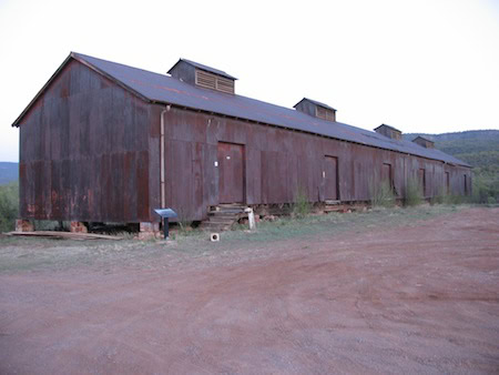 A long, weathered, brown metal building with several doors and small roof vents sits on a dirt lot with sparse vegetation.