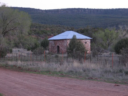 A small, square stone building with a metal roof sits near a dirt road, surrounded by trees and brush, with hills in the background.