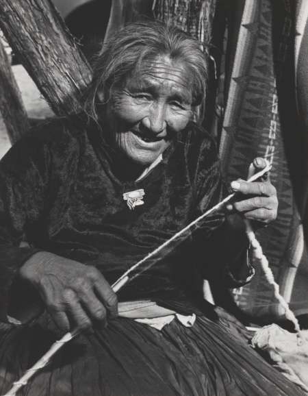 Elderly woman seated outdoors, smiling while spinning yarn by hand, with wooden posts and patterned textiles in the background.