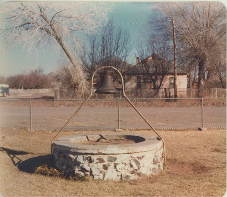 A stone well with a large metal bell mounted above it stands in a grassy yard near a road, with houses and bare trees in the background.