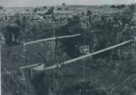 A black and white photo shows an old, makeshift wooden bridge spanning a rocky ravine in a rural landscape with sparse vegetation.
