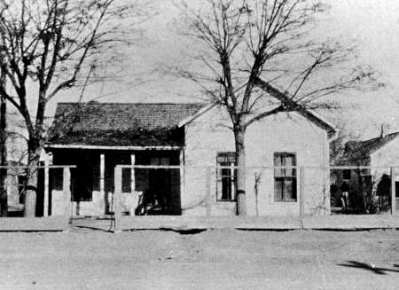 Black and white photo of a small, single-story house with a front porch, bare trees in front, and a wooden fence along the sidewalk.