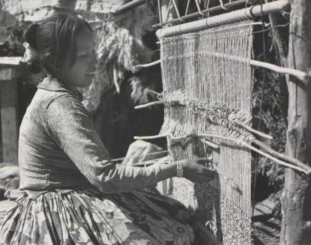 A woman sits outdoors, weaving fabric on a vertical loom using her hands. Various threads and tools are visible around her.
